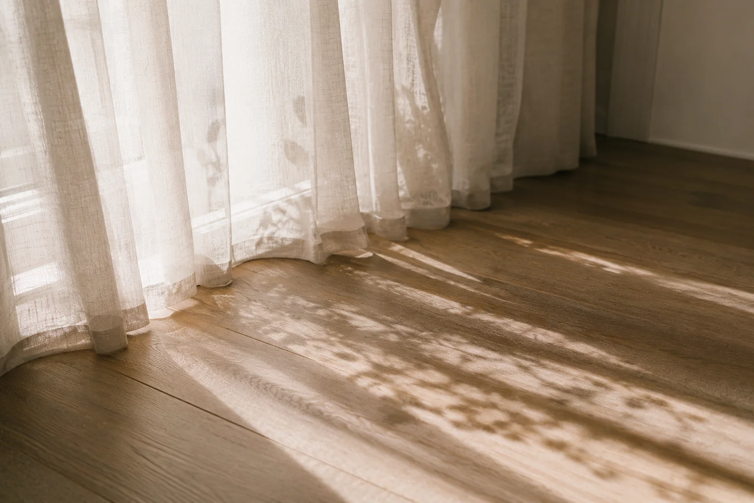 Sheer white curtains in a sunlit room with dappled leaf shadows across warm timber floors